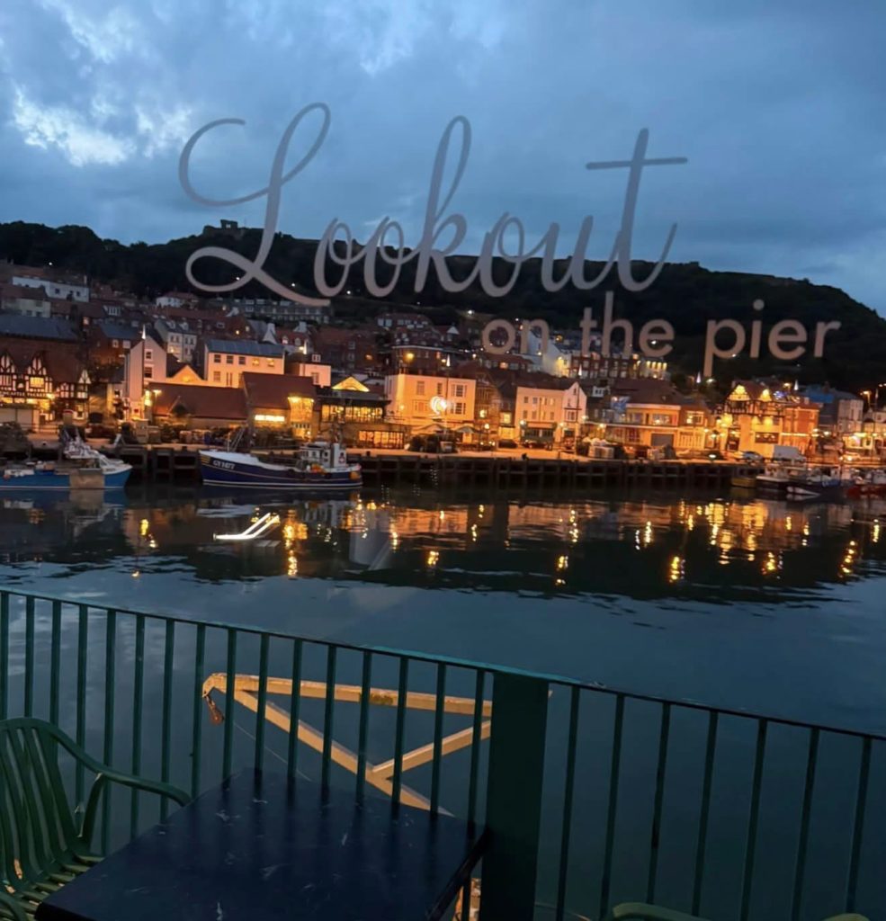 Evening view over Scarborough harbour with lit buildings and boats, with the words “Lookout on the Pier” across the image