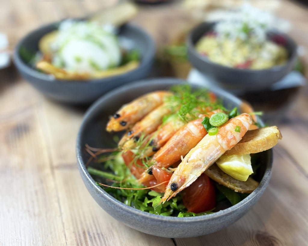 Prawns with lemon, tomato and rocket in a dark bowl, with blurred dishes behind